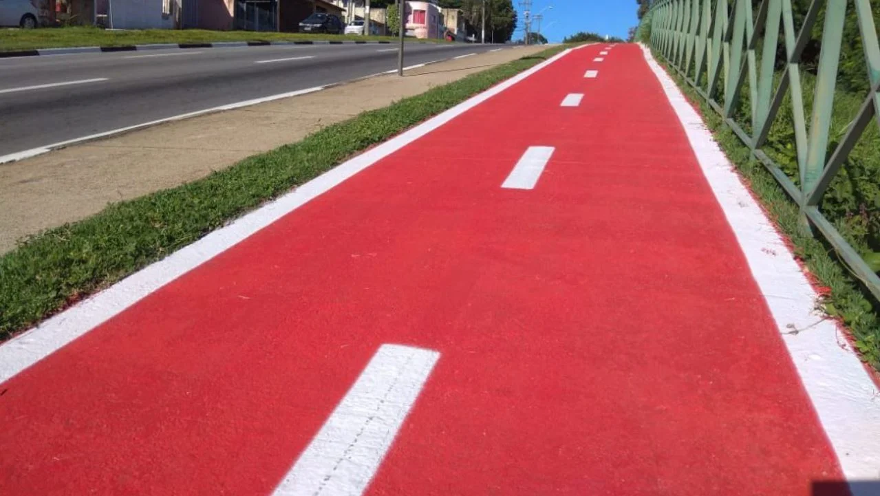 Foto mostra o piso de uma ciclovia, pintada em vermelho com faixas brancas