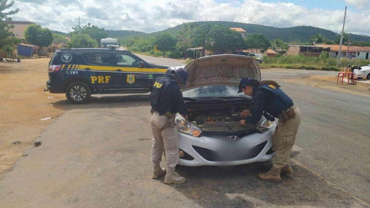 Foto mostra dois policiais vistoriando o veículo apreendido. Ao fundo, uma viatura da PRF