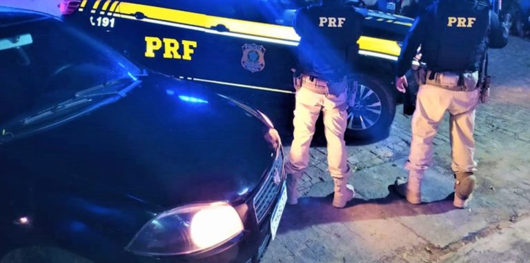 Imagem mostra parte da frente do veículo recuperado. Ao fundo, dois policiais rodoviários à frente de uma viatura da PRF