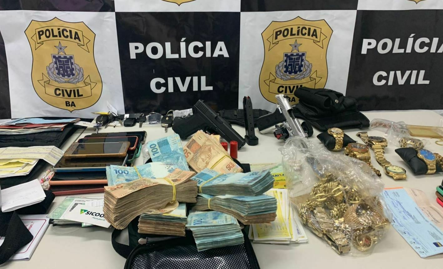 Foto mostra grande quantidade de dinheiro, celulares, armas, relógios, cheque e joias apreendidos em acampamento cigano. Ao fundo, um painel da Polícia Civil da Bahia