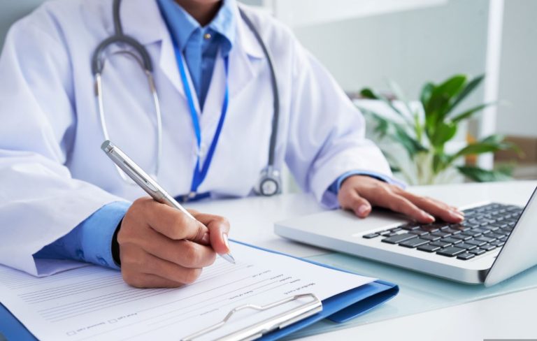 Foto mostra um homem vestido como médico usando um notebook e segurando uma caneta sobre uma prancheta