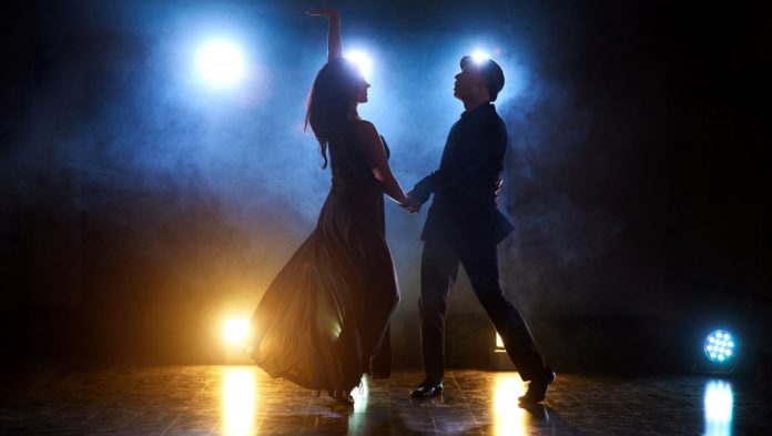 Foto mostra uma mulher e um homem dançando sobre um tablado e luzes de fundo como em um palco de teatro