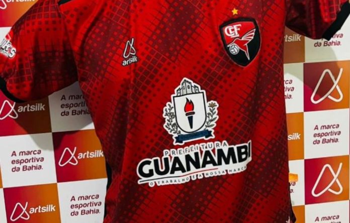 Foto mostra camisa do Flamengo de Guanambi, com cor predominantemente vermelha, com detalhes quadriculados preto e o brasão da Prefeitura de Guanambi como patrocinador principal. Ao fundo, um banner com a logomarca da Artsilk, fabricante dos uniformes