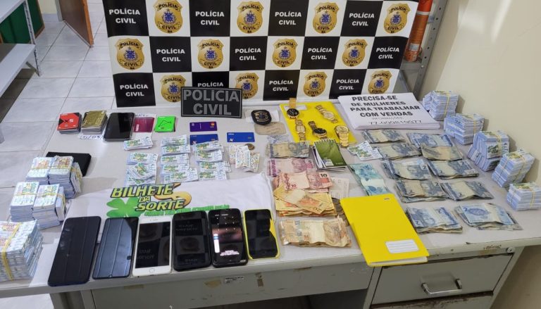 Foto mostra bilhetes de loterias, dinheiro, celulares, relógios e cartões de crédito. Ao fundo, um banner da Polícia Civil
