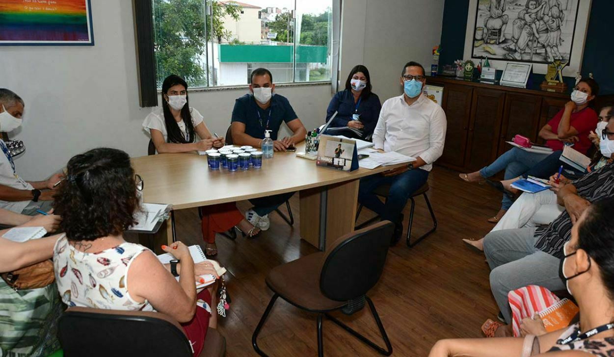 A imagem mosta a fotografia dos participantes da reunião que definiu a realização da Jornada da Cidadania. Os participantes estão sendatos em volta de uma mes, todos usam máscaras. Alguns fazem anotações em cadernos.