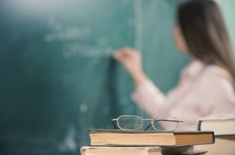 Foto desfocada mostra uma professora escrevendo em um quadro negro. No primeiro plano, alguns livros