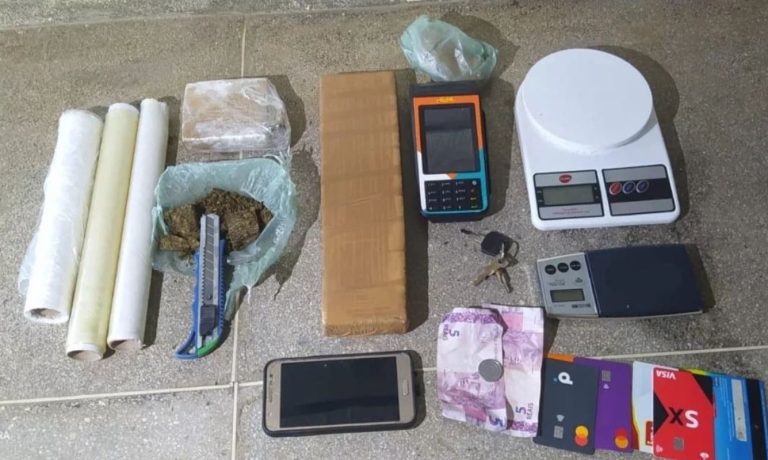 Foto mostra barra de maconha, embalagens, cartões bancários, uma máquina de cartão, um celular, uma quanta em dinheiro e duas balanças de precisão