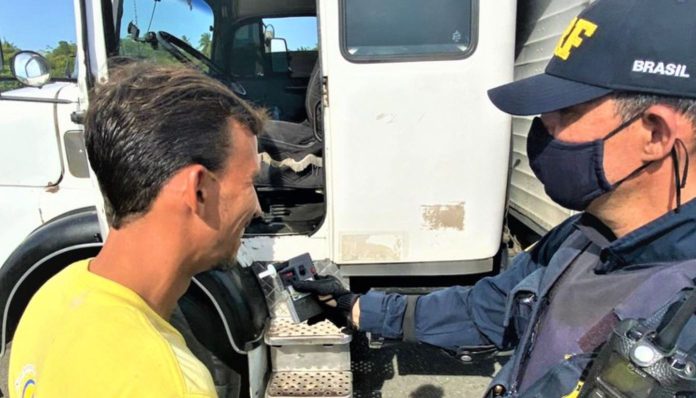 Foto mostra um policial rodoviário federal submetendo um caminhoneiro ao teste do bafômetro. A cabine do caminhão aparece ao fundo