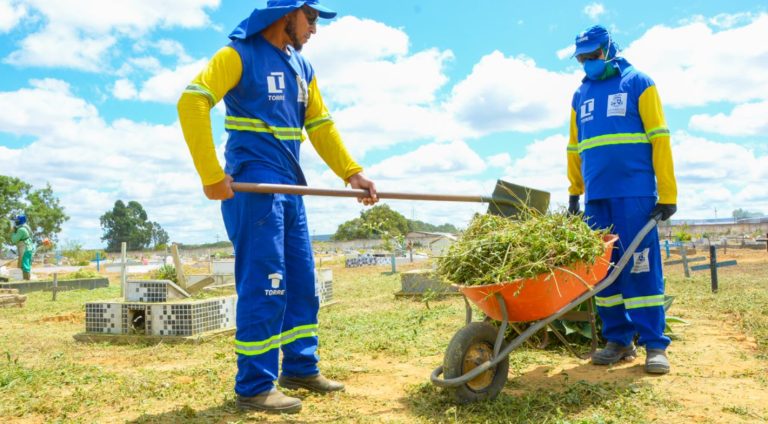Foto mostra um homem com uma pá e outro com um carrinho de mão sendo enchido de mato da limpeza do cemitério. Ao fundo é possível ver alguns túmulos.