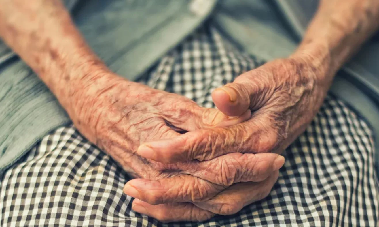 Foto mostra as mãos de uma idosa entrelaçadas sobre o colo