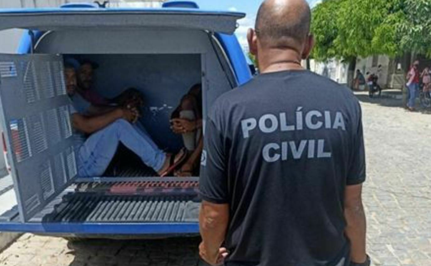 Foto mostra policial civil de costas e ao fundo homens presos dentro do compartilhamento da viatura